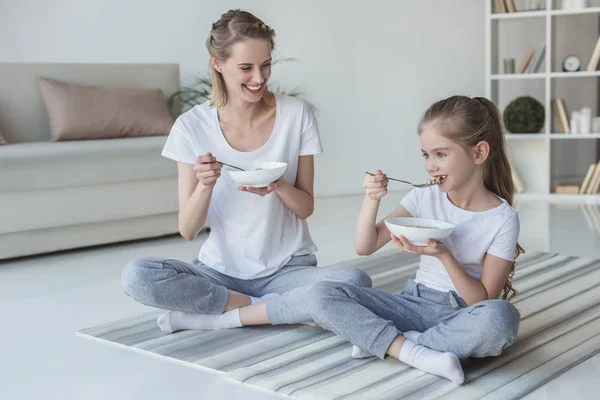 妈妈和女儿坐在瑜伽垫上吃谷类食物 — 图库照片