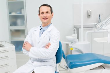 Gülümseyen doktor modern diş kliniğinde kollarını kavuşturarak oturuyor.