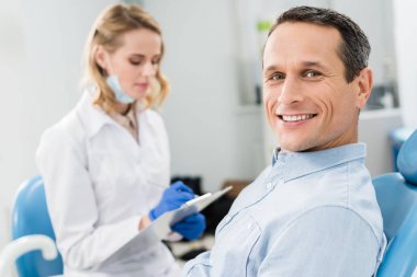 Dentist recording diagnosis while male patient waiting in modern clinic clipart