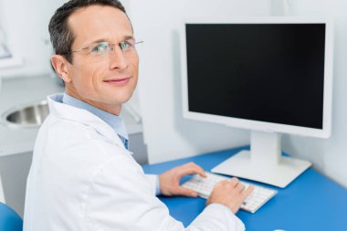 Modern klinik bilgisayar tarafından çalışma gözlük gülümseyen doktor