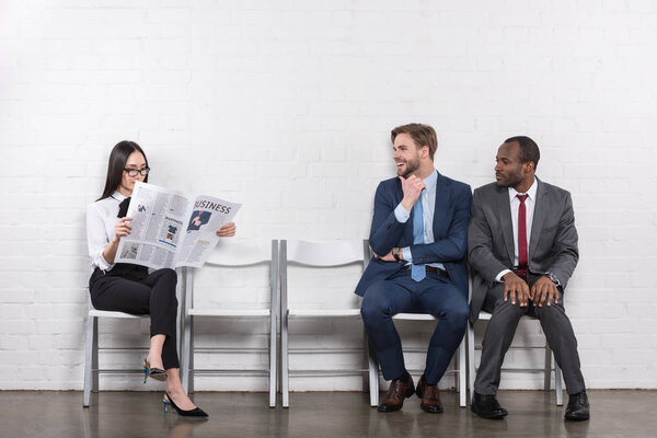 multicultural businessmen looking at asian businesswoman with newspaper while waiting for job interview