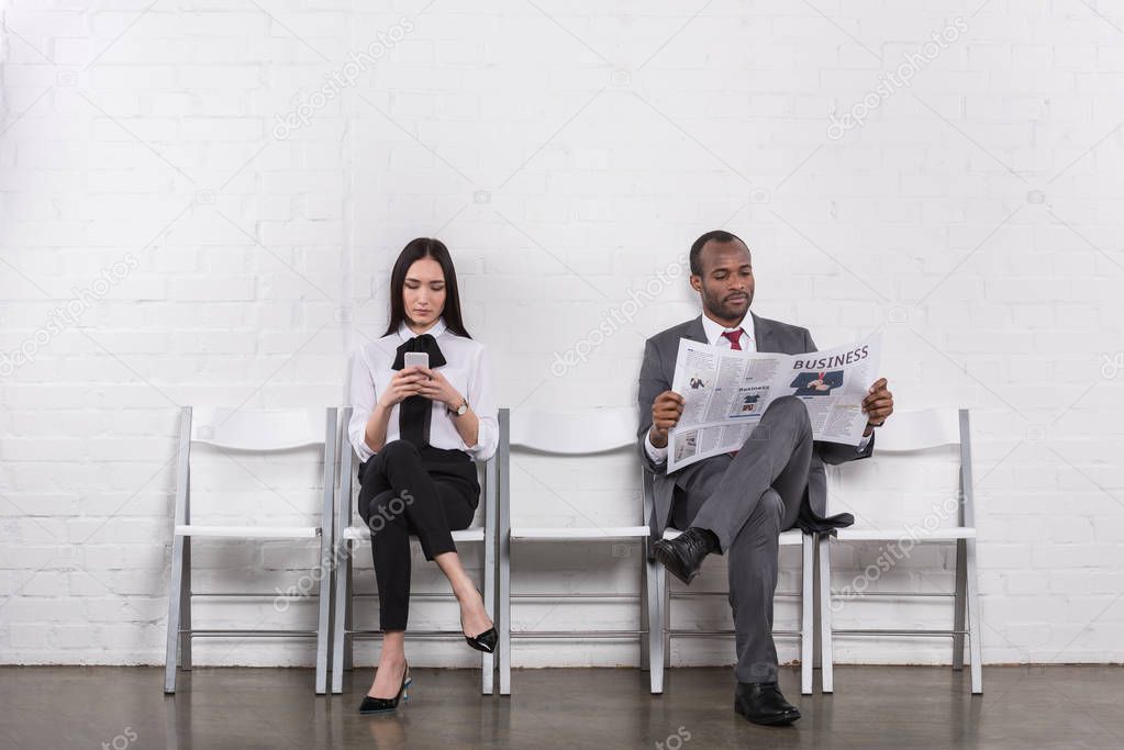 asian businesswoman with smartphone and african american businessman with newspaper waiting for job interview 