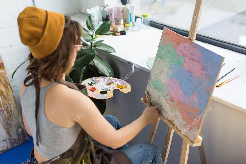 Young artistic girl working with painting knife and canvas in light studio