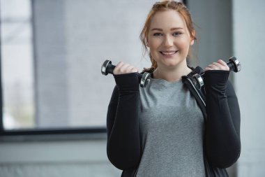 Obese smiling girl training with dumbbells in gym