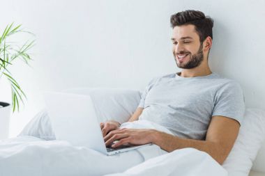 yatakta yatarken ve dizüstü bilgisayar kullanarak gülümseyen adam