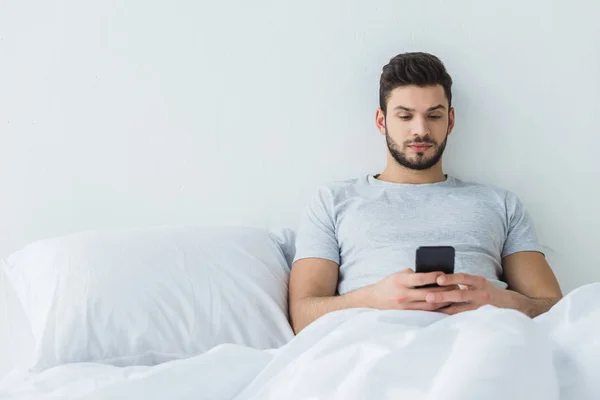 Бородатый Человек Лежит Кровати Отправляет Сообщения Смартфоне — стоковое фото