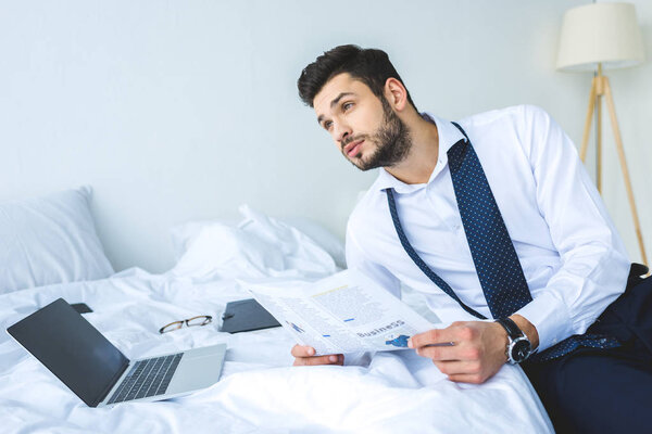 thoughtful businessman working with newspaper and laptop on bed in morning