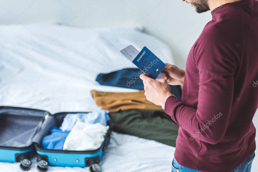 cropped view of man holding passport and ticket, suitcase with clothes on background