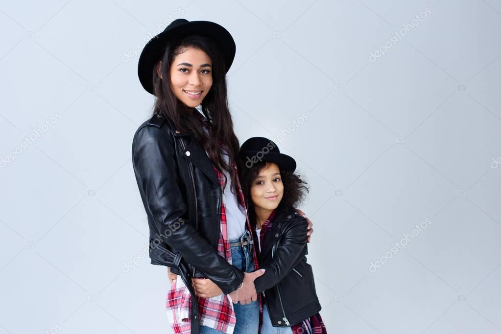 stylish beautiful mother and daughter in similar clothes embracing and looking at camera isolated on grey