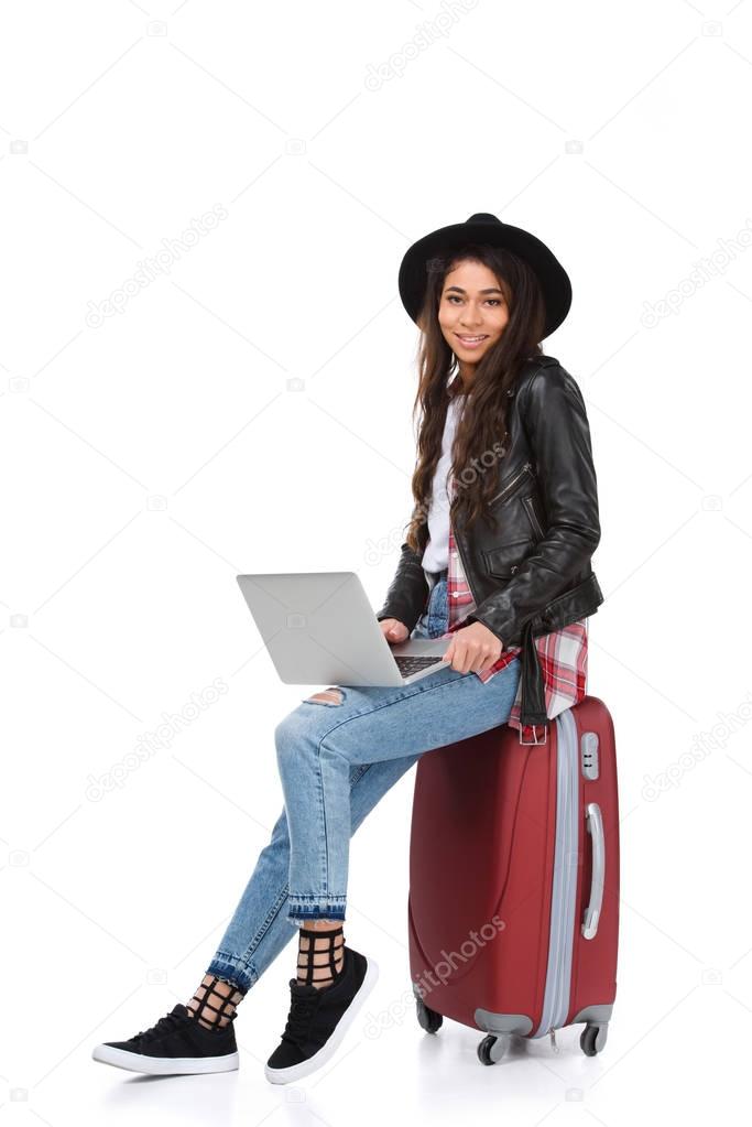 stylish young woman using laptop while sitting on luggage isolated on white