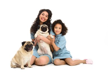 happy young mother and daughter sitting on floor with two adorable pugs isolated on white clipart