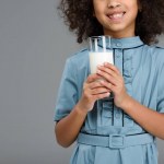 Abgeschnittene Aufnahme eines lächelnden kleinen Kindes mit einem Glas Milch isoliert auf grau