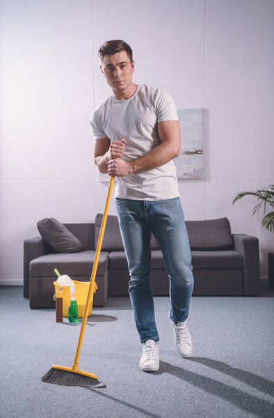 handsome man sweeping living room with broom and looking at camera