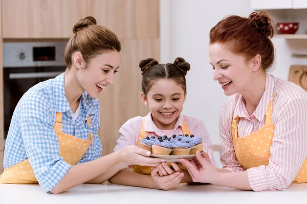 three generations of women in aprons with delicious cupcakes on tray at kitchen