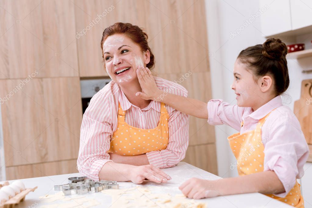happy grandmother and little granddaughter playing with flour at kitchen while cooking