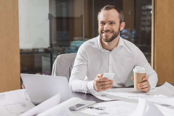 happy architect with paper cup of coffee using smartphone at office