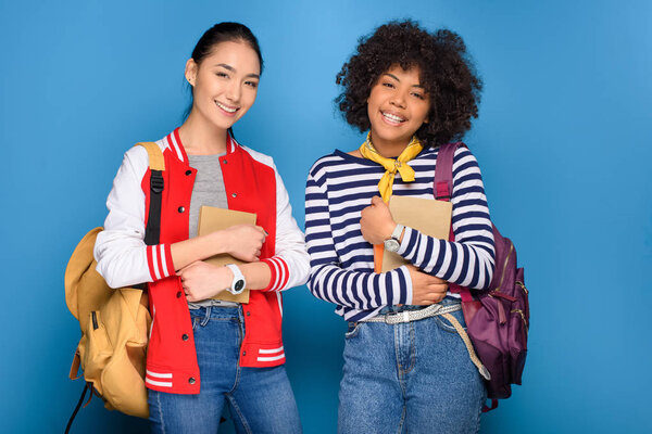 happy female african american and asian students posing with books, isolated on blue