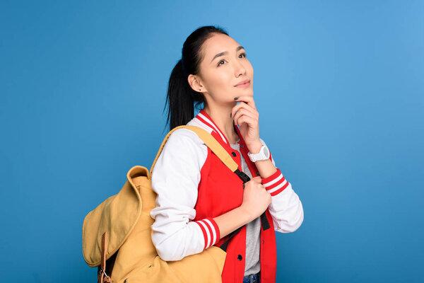 side view of pensive asian student with backpack isolated on blue