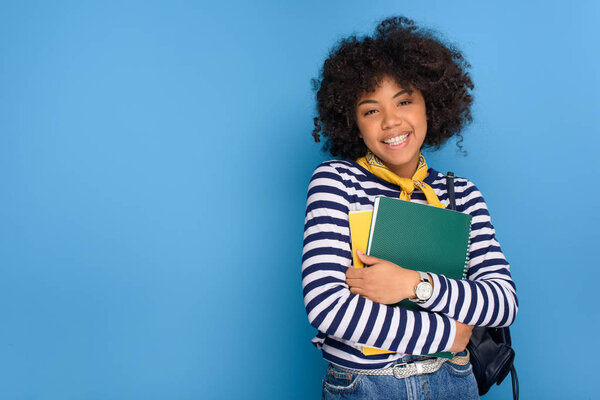 portrait of cheerful african american student with notebooks isolated on blue