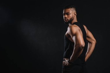 handsome young african american sportsman standing with hands on waist and looking at camera isolated on black clipart