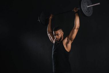 muscular young african american sportsman lifting barbell and looking away isolated on black clipart