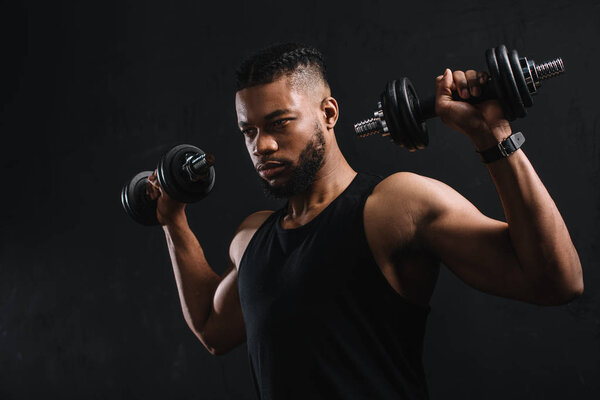 концентрированный африканский американский спортсмен, тренирующийся с гантелями, изолированными на черном
 