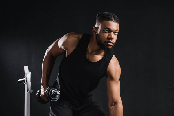 young african american sportsman exercising with dumbbell and looking away on black