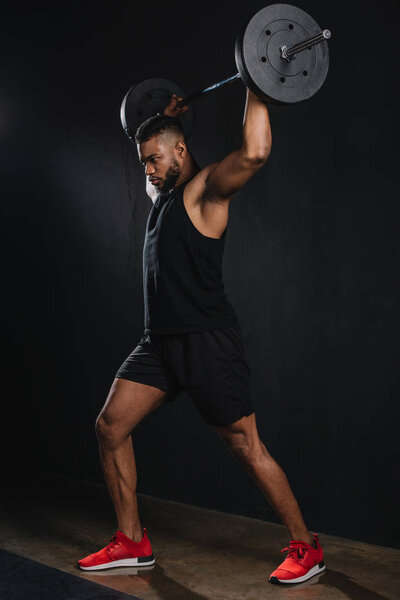 full length view of muscular young african american sportsman lifting barbell on black 