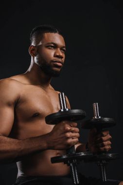 muscular shirtless young african american man holding dumbbells and looking away on black clipart