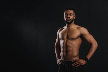 muscular shirtless african american man standing with hands on waist and looking at camera isolated on black clipart