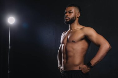 muscular young african american sportsman standing with hands on waist and looking away on black clipart