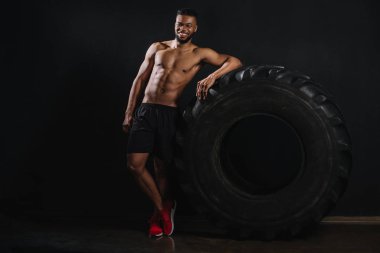 Lastik yaslanmış ve siyah kameraya gülümseyen kas gömleksiz genç Afro-Amerikan sporcu