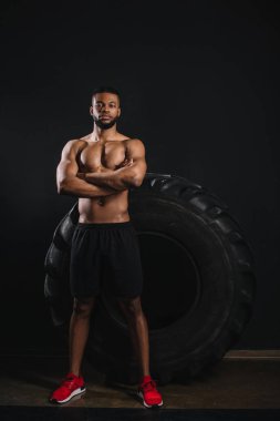 büyük lastik yakın çapraz kollar ile ayakta ve siyah kameraya bakarak gömleksiz Afrikalı-Amerikalı sporcu görünümünü tam uzunlukta 