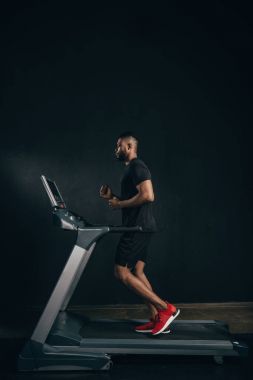 side view of young african american sportsman running on treadmill on black clipart