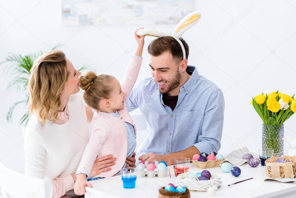 Child and parents playing with bunny ears by table with Easter eggs