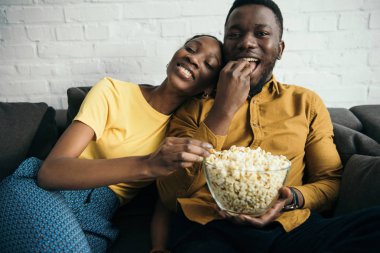 güzel mutlu Afrika kökenli Amerikalı çift evde kanepede otururken patlamış mısır yemek  