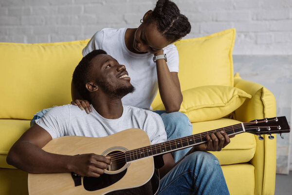 handsome smiling young man playing guitar and looking at beautiful girlfriend at home