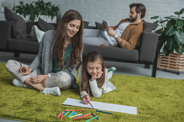 mother and daughter drawing on floor and father using tablet on sofa 