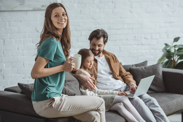father and daughter watching something at laptop and mother holding cup of coffee