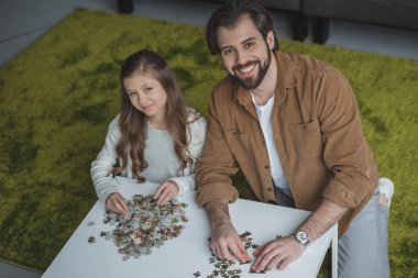 Baba ve kızı puzzle parçaları ile masada oturan ve kameraya bakarak yüksek açılı görünüş