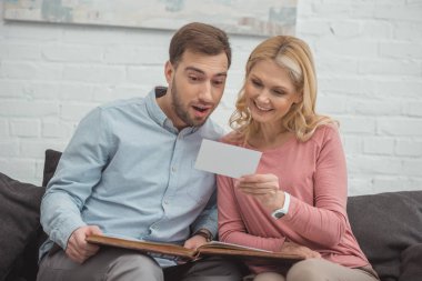 mother and grown son looking at photo in hand while resting on sofa together clipart