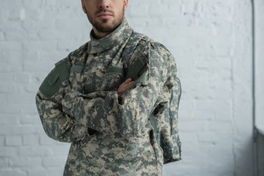cropped shot of soldier in military uniform with arms crossed standing against white brick wall clipart