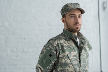 portrait of soldier in military uniform looking at camera against white brick wall clipart