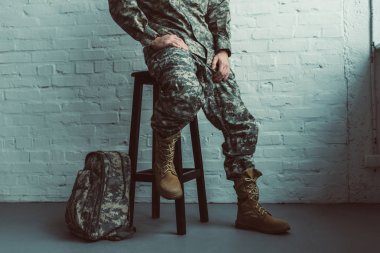 partial view of soldier in military uniform sitting on chair against white brick wall clipart
