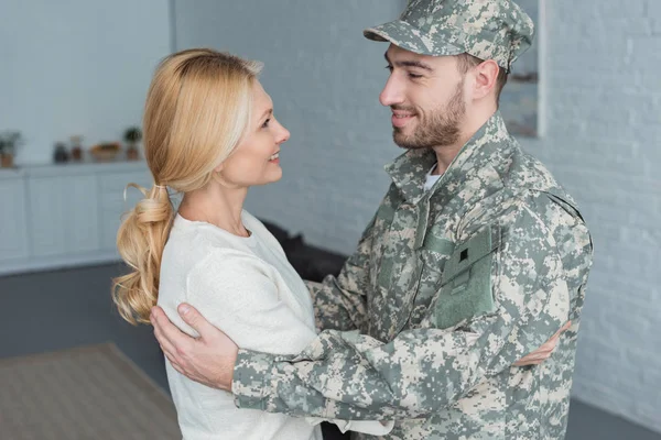side view of smiling man in military uniform and mother hugging each other at home