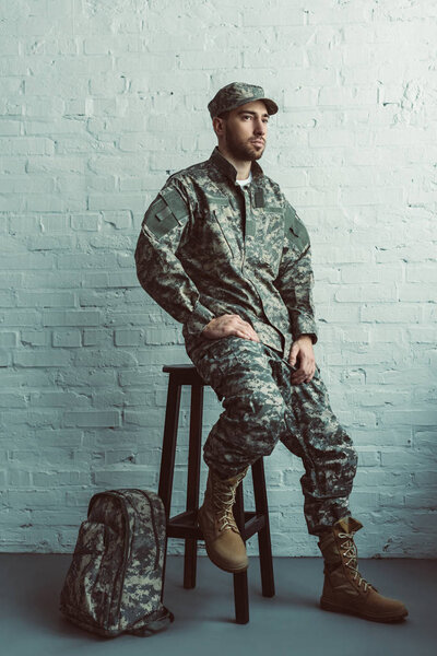 pensive soldier in military uniform sitting on chair against white brick wall