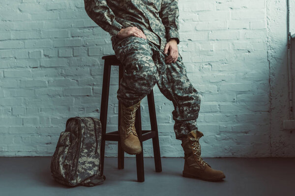 partial view of soldier in military uniform sitting on chair against white brick wall