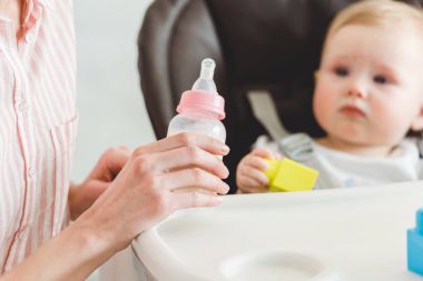 Besleme şişe ve plastik bloklarla bebek sandalyede oturan bebek kızı tutan anne görünümünü kırpılmış