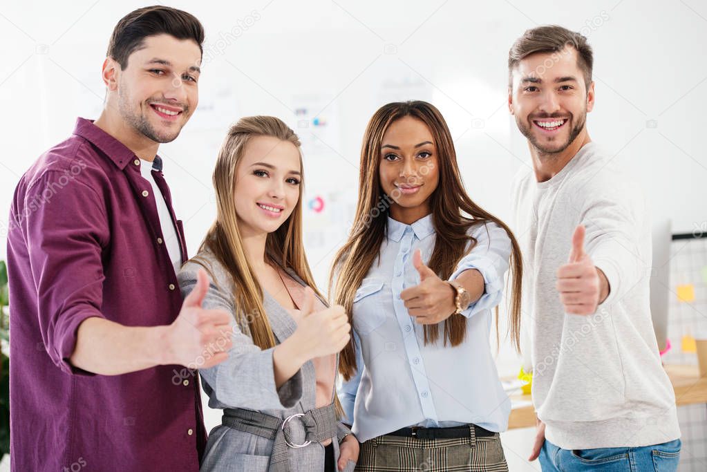 portrait of young smiling multiracial business people showing thumbs up and looking at camera in office