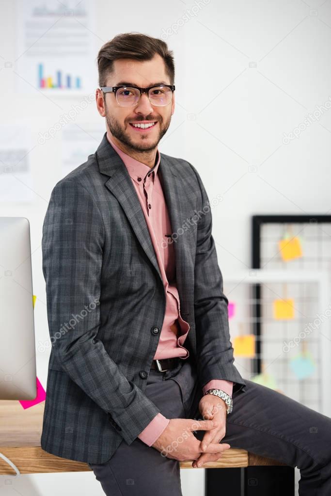 portrait of smiling businessman in suit and eyeglasses sitting on table and looking at camera in office